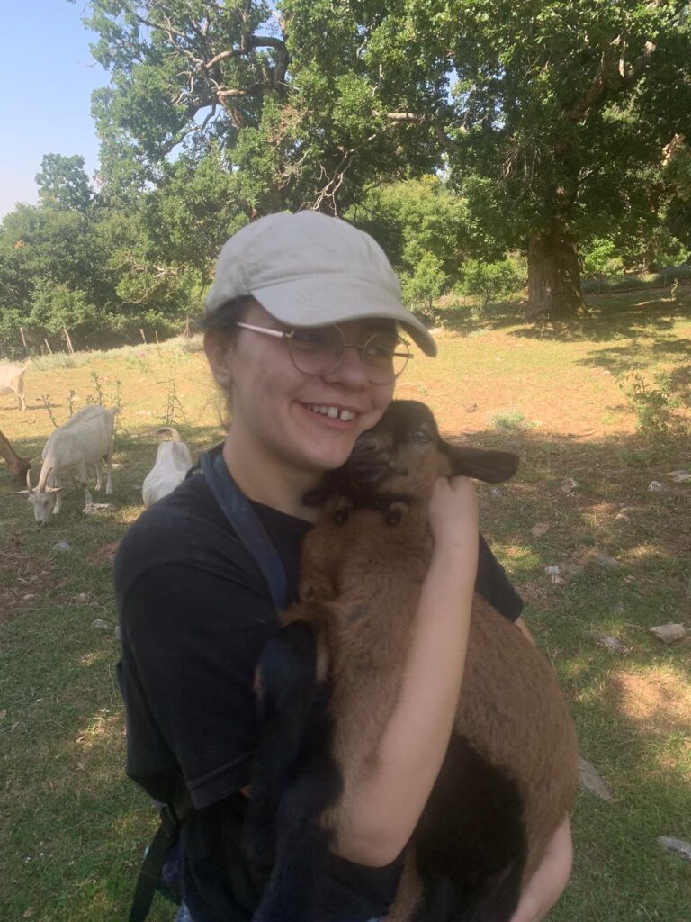 Lena Wiebe is pictured holding a brown and black baby goat. She is smiling in front of the background of a field and other white goats.